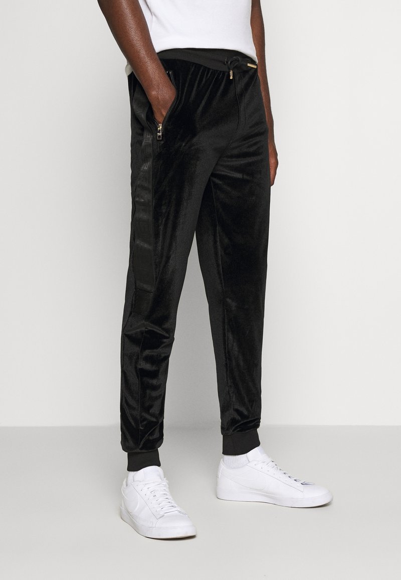 Eine Person steht da und trägt schwarze Samtjogginghosen mit seitlichen Reißverschlusstaschen und weißen Sneakers, was ein lässiges, stilvolles Outfit zeigt.