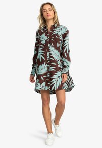 Brown shirt dress with light blue palm leaf pattern, long sleeves, button-down front, and a relaxed fit; paired with white sneakers.
