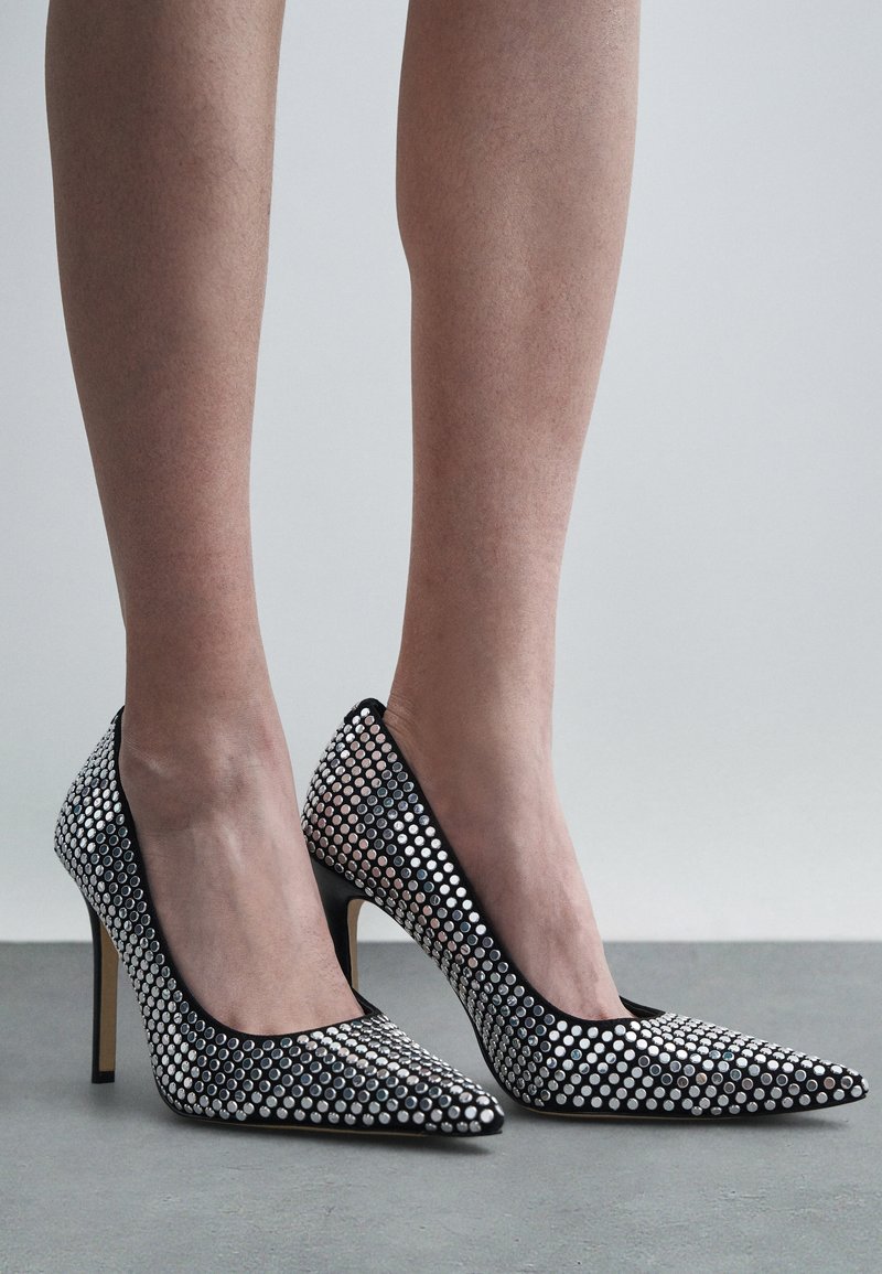 Black stiletto heels adorned with silver circular studs, featuring a pointed toe and smooth texture. The heels are high and slender.