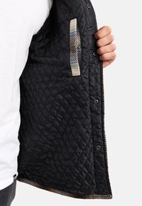 Interno di giacca trapuntata nera con una tasca testurizzata che presenta un motivo a quadri. Chiusure a scatto sul lato sinistro, fodera liscia.