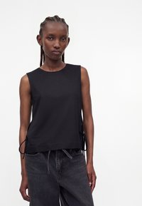 Woman with braided hair wearing a sleeveless black top with side ties and black jeans, standing against a plain white background.