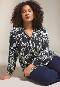 Marinebluse mit weißen abstrakten Mustern, gerafftem Ausschnitt, Bindebändern am Kragen, langen Ärmeln, strukturiertem Stoff, kombiniert mit dunkelblauen Hosen.