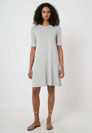 Kobieta o kręconych włosach, ubrana w jasnoszarą sukienkę z krótkim rękawem sięgającą kolan oraz beżowe, płaskie sandały na paski, stojąca na białym tle.