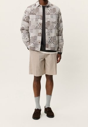 Mand iført en mønstret lys jakke, mørk skjorte under, beige knælange shorts, grå sokker og mørkebrune sko, stående med hænderne i lommerne.