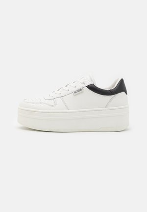 Basket à plateforme blanche avec détail noir au talon, lacets blancs et étiquette de marque Guess sur le côté, sur fond uni.