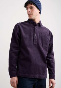 Purpursarkans samta audumā ar pogu apkakli, krūškurvi un teksturētu virsmu, savienots ar tumšsīkrēmu džinsiem un tumši zilā cepurē.