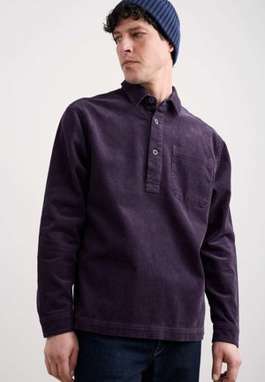 Lila Cord-Hemd mit Knopfkragen, Brusttasche und strukturierter Oberfläche, kombiniert mit dunkelblauen Denim-Jeans und einer marinefarbenen Mütze.