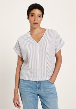 Junge Frau mit kurzen lockigen Haaren, die eine weiße Kurzarmbluse mit V-Ausschnitt und dünnen blauen Streifen sowie helle blaue Jeans trägt, stehend mit einer Hand in der Tasche.