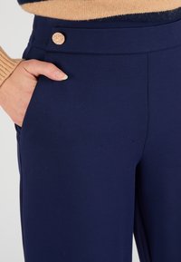 Marineblauwe broeken met een gladde textuur, voorzien van een goudkleurige decoratieve knoop bij de taille en zijzakdetails.
