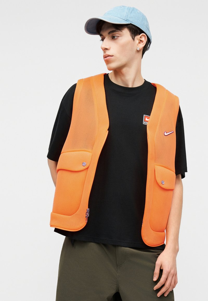 Junger Mann mit hellblauer Kappe, schwarzem Nike-T-Shirt und leuchtend orangefarbener Weste mit großen Taschen, der seitlich vor einem schlichten weißen Hintergrund schaut.