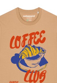 Beige T-shirt met een blauwe illustratie van een koffiekop en croissant, voorzien van rode tekst "COFFEE CLUB". Gemaakt van een katoenen mix, met een ronde hals.