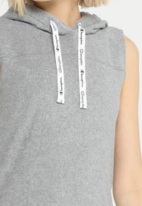 Szary bezrękawnik z miękkiego materiału, z przednią kieszenią i białymi sznurkami do ściągania z czarnymi logo tekstowymi.