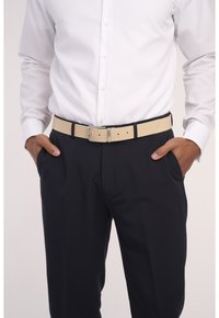 Beiger perforierter Leder-Gürtel mit einer silbernen Metallschnalle, getragen über dunklen Anzughosen. Weißes, kurzärmeliges Hemd ist in die Hose gesteckt.