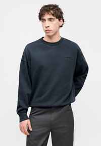 Σκούρο μπλε φούτερ από απαλό ύφασμα, με στρογγυλή λαιμόκοψη, χαμηλωμένους ώμους και διακριτικό λογότυπο μπροστά. Συνδυάζεται με γκρι παντελόνι.