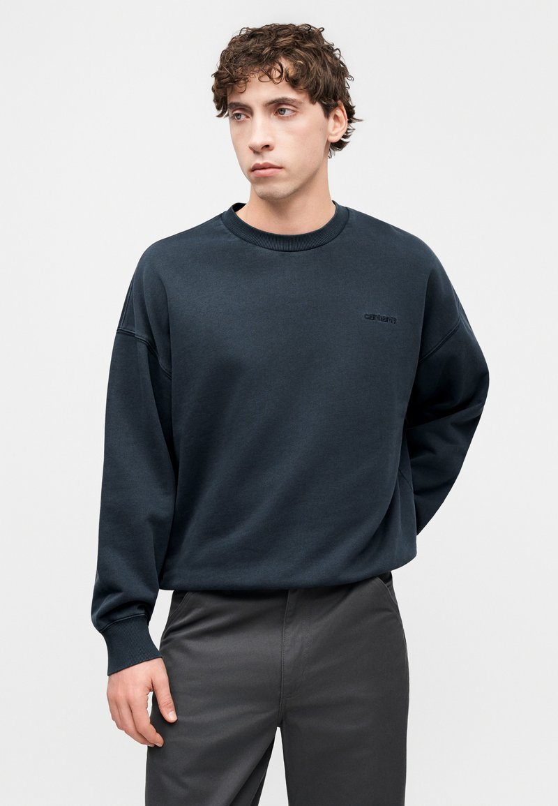 Felpa blu scuro in tessuto morbido, con collo rotondo, spalle a sbuffo e un logo discreto sul davanti. Abbinata a pantaloni grigi.