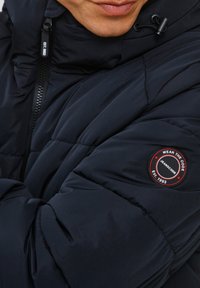 Granatowa kurtka puffer z pikowanym wykończeniem, z suwakiem i okrągłą łatą z logo na lewym rękawie.