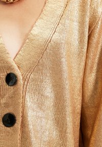 Goldener Strickcardigan mit V-Ausschnitt, versehen mit schwarzen Knöpfen und einer glatten, strukturierten Oberfläche.