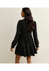 Vestido negro en capas con mangas largas y abullonadas. Tejido texturizado, dobladillo con volantes, corsé ajustado. Los detalles incluyen puños fruncidos para un diseño adicional.