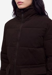 Femme portant une veste matelassée marron foncé avec fermeture éclair, col montant et poches à rabat, vue des épaules au menton.