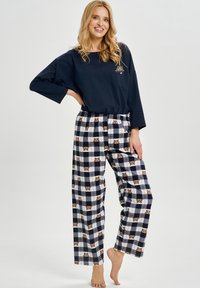 Granatowa bluzka z długim rękawem z nadrukiem niedźwiedzia, zestawiona z flanelowymi spodniami do piżamy w kratę z motywem niedźwiedzi w kolorach brązowym, granatowym i białym. Miękki materiał.