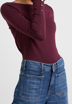 Vrouw draagt een aangesloten bordeauxrode top met gouden knopen op de mouwen en een hoog getailleerde blauwe spijkerbroek met grote voorkantzakken.