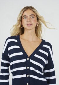 Blonde vrouw met los haar draagt gouden creolen en een donkerblauwe cardigan met brede witte horizontale strepen en gouden knopen.