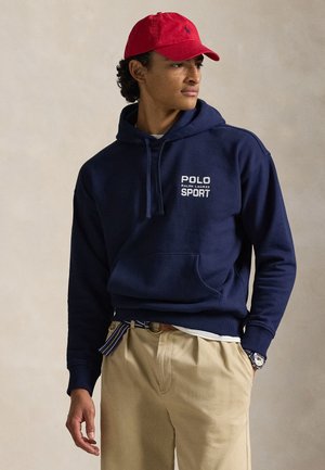 Sudadera con capucha azul marino con texto "POLO RALPH LAUREN SPORT". Cuenta con un bolsillo tipo canguro y capucha con cordón. Combinada con pantalones caqui y una gorra roja.