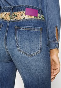 Calças de ganga azul-escuro com um padrão floral na cintura, uma etiqueta de marca roxa e bolsos traseiros clássicos com detalhes de costura.