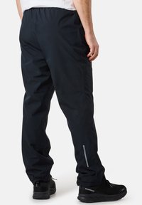 Schwarze wasserdichte Hose mit lockerer Passform, elastischem Bund, Reißverschlüssen an den Taschen und reflektierenden Akzenten an den Seiten.