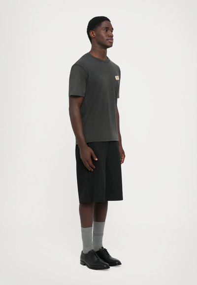 Jovem de pé, a usar uma t-shirt preta desbotada, calções pretos até aos joelhos, meias cinzentas e sapatos pretos de vestir, contra um fundo simples.