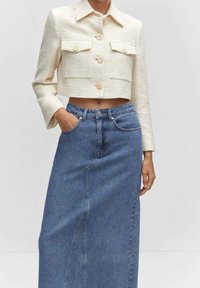 Chaqueta corta de cremoso con dos bolsillos frontales y botones, combinada con una falda de mezclilla azul de tiro alto. Tela texturizada y diseño clásico.