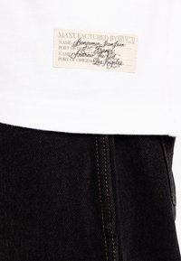 White cotton t-shirt with a small beige label sewn on, featuring black text. Paired with dark denim jeans with yellow stitching.