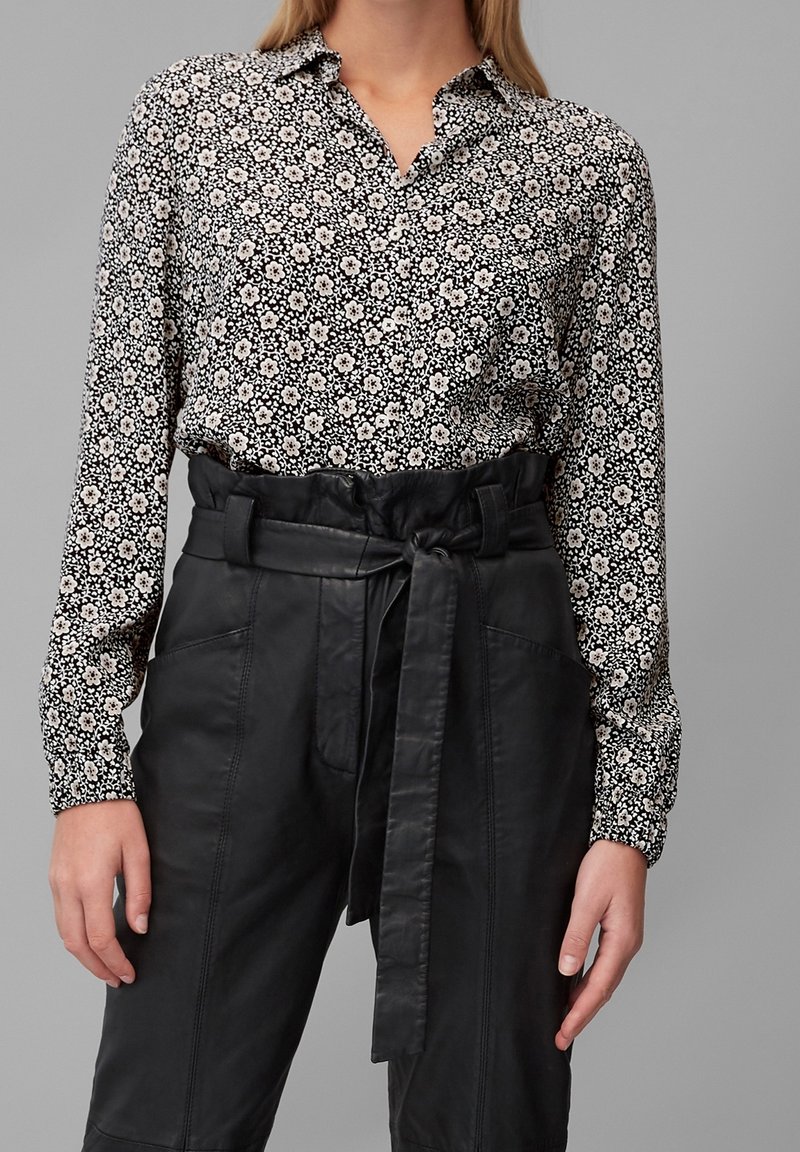 Femme portant un chemisier à motifs floraux noirs et blancs, boutonné et rentré dans un pantalon en cuir noir taille haute avec une ceinture nouée.