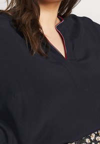 Navyblauwe blouse met een V-halsontwerp en een rood-witte afwerking. Soepele stof, iets losvallend model, met een klein zakje.