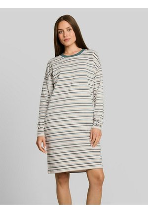 Jonge vrouw die een jurk met lange mouwen tot op de knie draagt met horizontale blauwe en beige strepen tegen een effen grijze achtergrond.