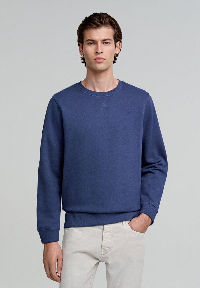 Marineblaues Sweatshirt mit Rundhalsausschnitt, gerippten Bündchen, strukturiertem Stoff und einem kleinen Logodetail auf der Brust, kombiniert mit hellen Hosen.