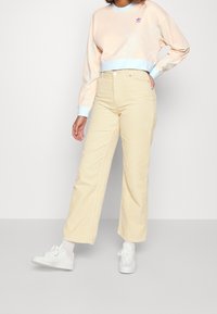 Felpa pesca chiaro con bordi blu e logo viola, abbinata a pantaloni a vita alta in velluto a coste gialli e sneakers bianche.