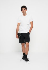 Mann som står og har på seg en hvit t-skjorte med logo, svarte shorts, svarte sokker, svarte joggesko, og holder en liten svart pose i venstre hånd.