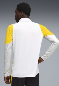 Camisa atlética de manga larga con un cuerpo blanco, hombros amarillos y mangas grises con patrón. Fabricada con un tejido ligero y texturizado.