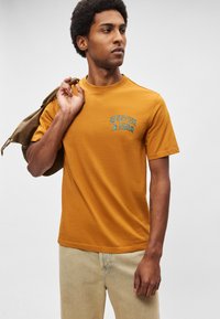 Μουσταρδί κίτρινη κοντομάνικη μπλούζα με στρογγυλή λαιμόκοψη, με μπλε κείμενο «SCOTCH & SODA» και λεία βαμβακερή υφή.