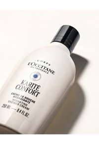 Vit plastflaska med svart lock. Etiketten visar svart och blå text som anger återfuktande duschkräm med sheasmör. 250 ml.