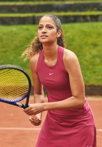 Atleta con un vestido rosa ajustado sin mangas y el logo de Nike, sosteniendo una raqueta de tenis con un patrón de cuerdas azul y amarillo en una cancha de tierra batida.