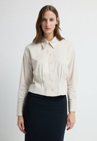 Kremowa bluzka zapinana na guziki z kołnierzykiem, długimi rękawami i marszczeniami w talii. Materiał wydaje się gładki i lekki.