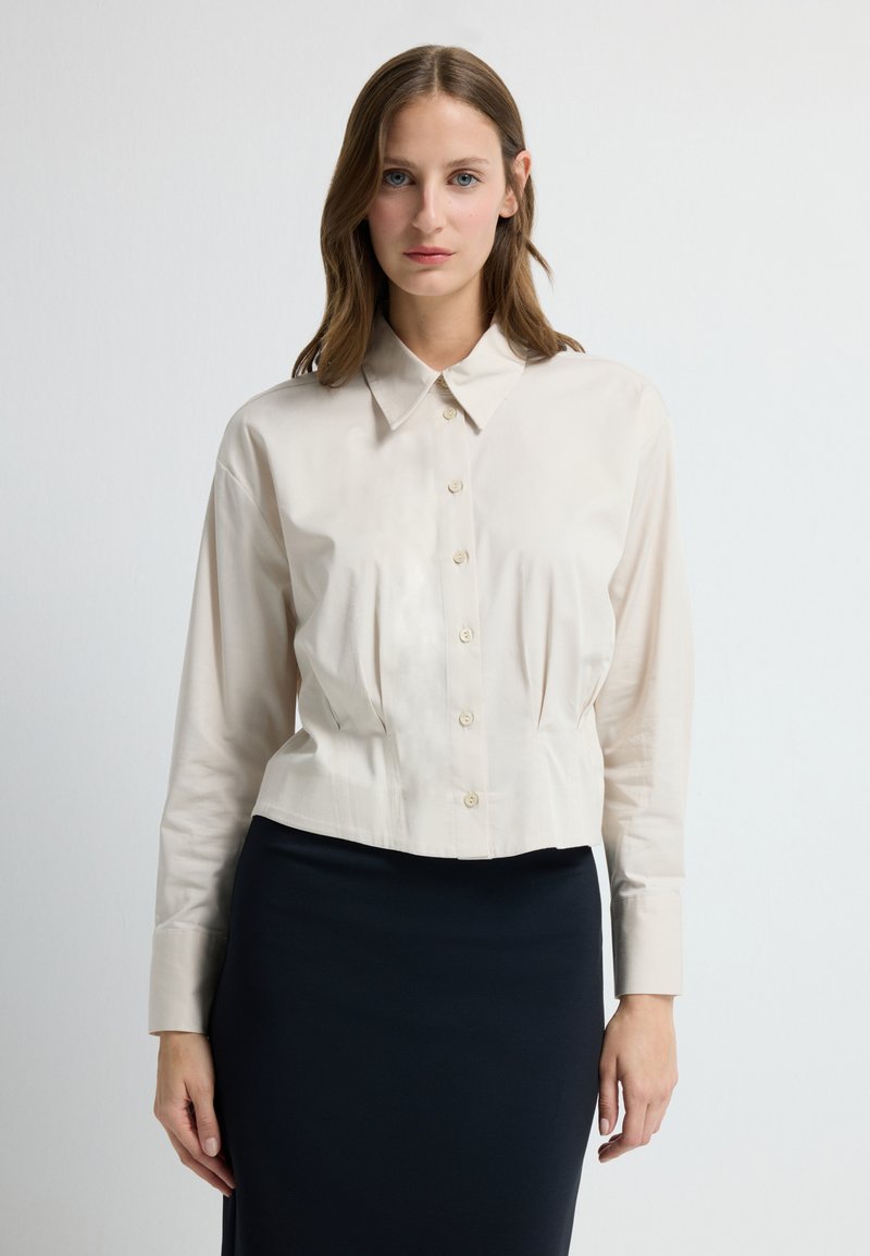 Kremowa bluzka zapinana na guziki z kołnierzykiem, długimi rękawami i marszczeniami w talii. Materiał wydaje się gładki i lekki.