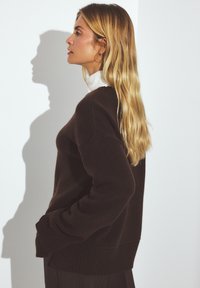 Ruskea oversized neulepaita, jossa on ribbauspäät ja helma, pyöreä kaulus ja sivutaskut, päällä valkoinen poolopaita.