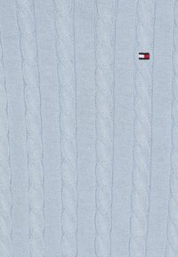 Lyseblå strikkegenser med kabelmønster, utstyrt med en liten brodert logo i rødt, hvitt og blått på forsiden.