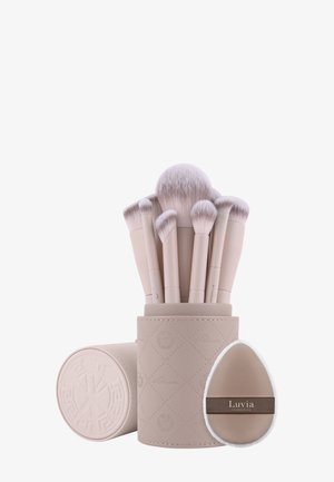 Conjunto de siete brochas de maquillaje beige con cerdas suaves en un estuche cilíndrico y una esponja de maquillaje beige etiquetada como "Luvia Cosmetics".