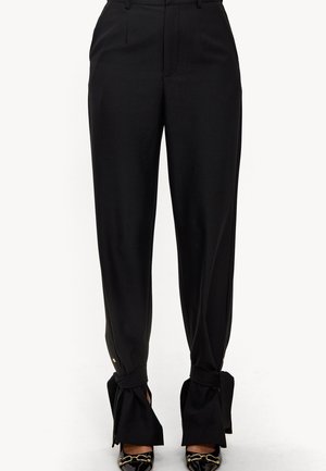 Zwarte hoge tailles broek met enkelstrikjes, gecombineerd met zwarte lakleren puntige schoenen met gouden kettinkdetail.