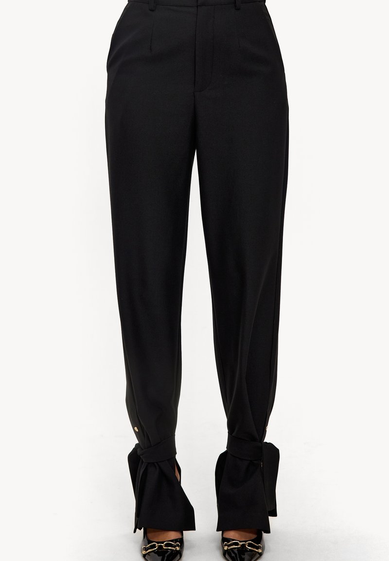 Pantalon noir taille haute avec liens à la cheville, associé à des chaussures pointues en cuir verni noir ornées d'un détail en chaîne dorée.