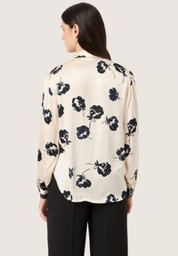 Long-sleeve blouse in cream fabric with black floral print, featuring a relaxed fit and button cuffs. Smooth texture and soft drape.